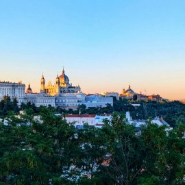 A blue sky and green trees frame a sunrise view of Madrid's cathedral and Royal Palace.